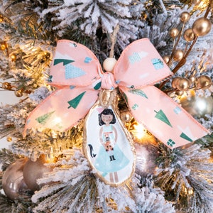 May include: A pink and green patterned ribbon bow with a wooden bead hangs from a gold painted oyster shell ornament. The ornament features a hand-painted illustration of a girl in a blue dress holding a cat.