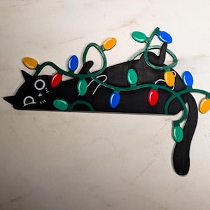 Pode incluir: Uma figura de gato preto emaranhada em luzes de Natal verdes com lâmpadas coloridas. O gato tem um rosto branco com olhos pretos e uma pequena boca.