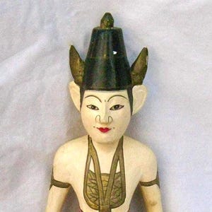 May include: A hand-painted wooden figurine of a seated figure with a black hat and large ears. The figure wears a green and red patterned skirt and a gold-colored necklace. The figurine is approximately 10 inches tall.