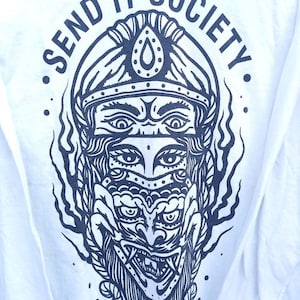 May include: White long-sleeve shirt featuring a detailed navy blue graphic of a multi-faced figure wearing a headdress. The text "SEND IT SOCIETY" is arched above the graphic, with small dots on either side.