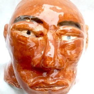 May include: A ceramic sculpture of a human head with a brown glaze. The head has prominent features, including a large nose, full lips, and defined eyebrows. The eyes are detailed with visible irises. The sculpture is set against a white background.