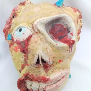 May include: A grotesque, handmade mask with a zombie theme. The mask is painted in shades of yellow, red, and blue, with details like an exposed eye, open mouth, and dripping blood. The mask is designed for Halloween or costume use.