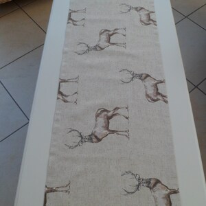 May include: A beige table runner featuring a repeating pattern of brown deer silhouettes. The runner is rectangular and appears to be made of a linen or similar fabric. The deer are depicted in a detailed, artistic style.