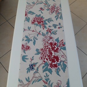 May include: A beige table runner with a floral pattern featuring large red and white flowers with teal green leaves.