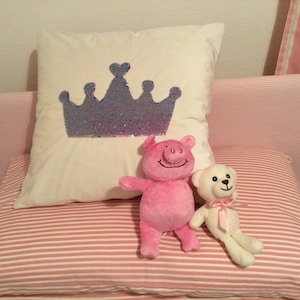 May include: A white pillow with a sequined crown design, a pink pig stuffed animal, and a white teddy bear with a pink bow. The pillows have a pink and white striped pattern. The scene is set on a bed.