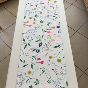 Op de afbeelding: Een witte tafelrunner met een bloemenpatroon met roze, blauwe, gele en groene bloemen. De runner ligt op een witte tafel.