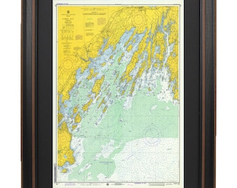 May include: A framed nautical chart of Casco Bay, Maine. The chart is printed on paper and features a yellow and green color scheme. The chart is framed in a black frame with a brown mat.