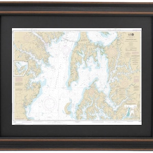 May include: Framed nautical chart of Chesapeake Bay, featuring detailed coastlines, waterways, and navigational markers. The map is printed on a cream-colored background with blue and tan details. The chart is framed in a black frame with a brown inner edge.