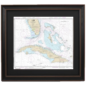 May include: A framed nautical chart of the Straits of Florida and Cuba. The chart is printed in black and white with blue and pink accents. The chart includes detailed information about the coastline, islands, and waterways. The chart is framed in a black frame with a brown mat.