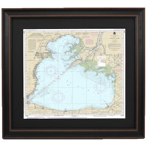 May include: A framed nautical chart of Lake St. Clair, showing depths, buoys, and other navigational features. The chart is printed on white paper with blue and green accents. The frame is black with a brown inner border.