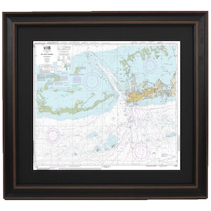May include: Framed nautical chart of Key West Harbor, featuring detailed water depth markings, islands, and navigational aids. The chart is in shades of blue, green, and tan, set within a black frame with a brown inner edge.