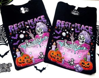 May include: Black t-shirts with a Halloween-themed graphic. The design features a skeleton in a bathtub with the words "REST IN PEACE" above it. Pumpkins, candles, and bats surround the skeleton. The brand logo "Better in Black co." is visible.