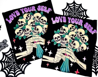 Può includere: Due magliette nere con la scritta "LOVE YOUR SELF" in viola. Ogni maglietta presenta una mano di scheletro che tiene un bouquet di teschi con foglie verdi. Il design include stelle e una ragnatela.