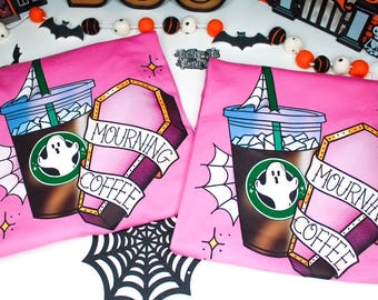 May include: Two pink t-shirts with a Halloween-themed design. Each shirt features a coffee cup with a ghost, a coffin with the words "MOURNING COFFEE", and a spiderweb. The design is in shades of brown, blue, and pink.