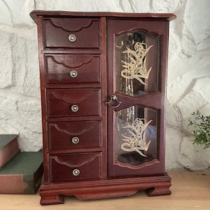 May include: A dark cherry-stained wooden jewelry armoire with six drawers and a mirrored door. The door features a gold floral design. Each drawer has a small, round silver knob. The armoire sits on a wooden shelf against a white stone wall.