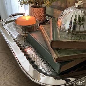 May include: A rectangular silver-toned metal tray with ornate embossed edges, holding two stacked green hardcover books, a small glass bowl with a lit orange candle, a small potted green plant, and a reflective silver-toned dome-shaped lid.