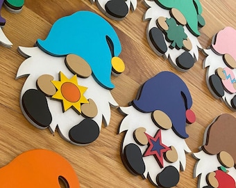 Puede incluir: Una colección de diez adornos de gnomo de madera, cada uno pintado en un color diferente y con un diseño único. Los gnomos sostienen varios artículos, incluyendo un sol, una estrella, un trébol, una calabaza, un pavo y un adorno navideño.