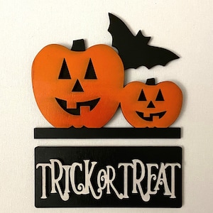 May include: A wooden Halloween sign with two orange jack-o'-lantern faces and a black bat. The sign says "Trick or Treat" in white letters.