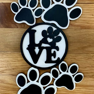 May include: Wooden paw print cutouts in black and white, along with a round sign that says "LOVE" with a paw print and heart. The paw prints are in various sizes and arranged on a wooden surface.