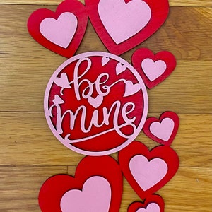 May include: Red and pink wooden hearts with the words "be mine" in a circle. The hearts are layered on top of each other and are arranged in a heart shape.