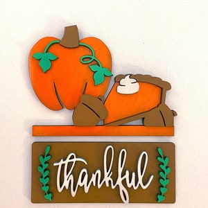 May include: Wooden fall-themed decor featuring an orange pumpkin with green vines, a slice of pumpkin pie, and two acorns. Below, a brown sign reads "thankful" in white script, with green leaf accents. Perfect for autumn decorating.