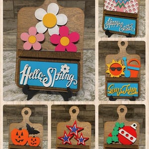 May include: Six wooden cutting boards with different seasonal designs and sayings. The boards have a small shelf and a stand. The designs include flowers, Easter eggs, a sun, pumpkins, stars, and a Christmas ornament. The sayings include "Hello Spring", "Happy Easter", "Sun & Fun", "Trick or Treat", "America", and "Merry & Bright".