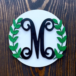 May include: A white round wooden sign with a black letter "M" surrounded by green leaves.