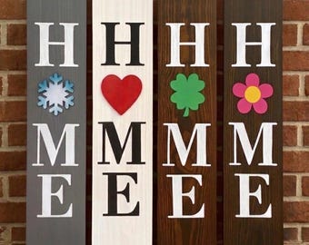 May include: Four vertical wooden signs with the word "HOME" in white and black lettering. Each sign features a different seasonal or holiday-themed accent: a snowflake, a heart, a shamrock, and a flower. The signs are displayed against a brick wall.