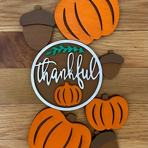 May include: A wooden craft project featuring the word "thankful" in white lettering on a brown circle with green leaves. The circle is surrounded by four orange pumpkins and two brown acorns.
