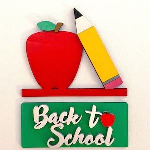 May include: Wooden sign with the words "Back to School" in white lettering on a green background. A red apple and a yellow pencil are above the sign.