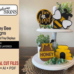 Puede incluir: Un expositor de bandejas de dos niveles blanco con cuatro decoraciones con temática de abejas. El nivel superior tiene una abeja amarilla y negra con alas blancas y un letrero redondo que dice "Bee kind" con una flor amarilla. El nivel inferior tiene un tarro de miel marrón con una etiqueta amarilla que dice "Honey" y un panal de abejas amarillo y negro.