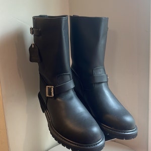 May include: A pair of black leather engineer boots with buckles. The boots are on a metal display stand.