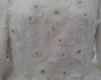 Puede incluir: Vestido de novia blanco con delicado bordado de encaje y adornos brillantes. El corpiño presenta un escote de corazón y la falda es amplia y fluida.