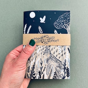 May include: A rectangular notebook featuring a dark blue night sky with a white moon and owl above a tan field with a hare and wheat stalks. A brown paper belly band wraps around the center, displaying a circular logo with the letters L and F.