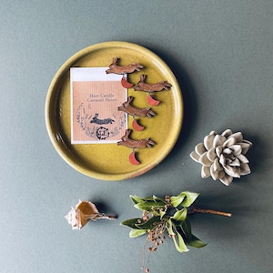 May include: Five wooden hare carousel pieces on a yellow plate. The hares are brown with red accents and are in a leaping pose. The text on the card reads "Hare Candle Carousel Pieces".