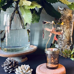 May include: A gold metal candle holder with a glass votive candle. The candle holder features a rotating carousel with a deer and other woodland creatures. The carousel is suspended from the top of the candle holder.