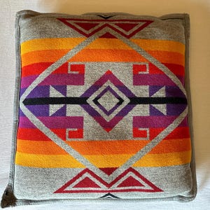 May include: Square cushion with a grey border and a vibrant geometric pattern. The pattern features a central diamond shape with red, orange, yellow, purple, and black accents. The cushion is likely made of a soft fabric.