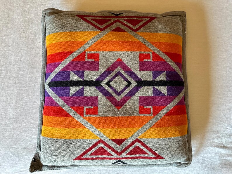 May include: Square cushion with a grey border and a vibrant geometric pattern. The pattern features a central diamond shape with red, orange, yellow, purple, and black accents. The cushion is likely made of a soft fabric.