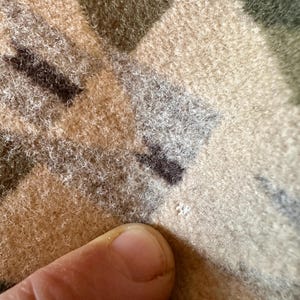 Może przedstawiać: Zbliżenie na teksturowaną tkaninę z wzorem kamuflażu w odcieniach beżu, brązu i zieleni. Tkanina wydaje się miękka i puszysta, z lekko zużytym wyglądem. Palec dotyka tkaniny.