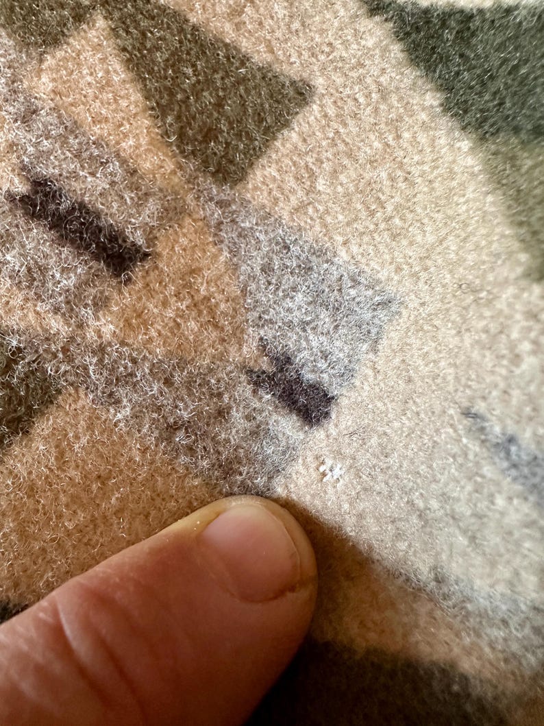 Może przedstawiać: Zbliżenie na teksturowaną tkaninę z wzorem kamuflażu w odcieniach beżu, brązu i zieleni. Tkanina wydaje się miękka i puszysta, z lekko zużytym wyglądem. Palec dotyka tkaniny.