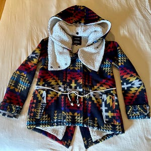 May include: A hooded jacket featuring a multi-colored geometric pattern in shades of red, orange, yellow, blue, and black. The garment includes a white sherpa fleece lining, a white drawstring waist, two front pockets, and long sleeves with cuffs.