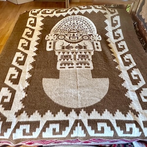 Peut inclure: Une couverture tissée rectangulaire marron et crème présentant une figure anthropomorphe stylisée centrale avec une grande coiffe. La bordure affiche un motif géométrique répétitif en escalier crème sur fond marron.