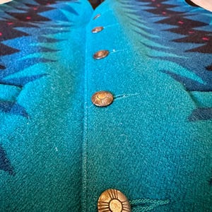 May include: A teal and blue patterned vest with four decorative buttons. The vest features a geometric design in shades of blue and black, with small red accents. The buttons are round and metallic, adding a vintage touch to the garment.