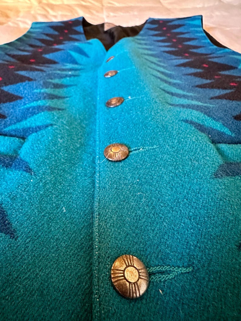 May include: A teal and blue patterned vest with four decorative buttons. The vest features a geometric design in shades of blue and black, with small red accents. The buttons are round and metallic, adding a vintage touch to the garment.