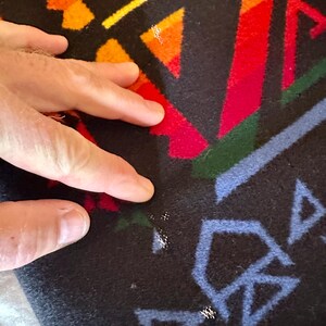 Puede incluir: Primer plano de un textil negro con un patr&oacute;n geom&eacute;trico en rojo, naranja, amarillo, verde y azul. El dise&ntilde;o presenta tri&aacute;ngulos y formas angulares. El textil tiene un aspecto suave y texturizado.