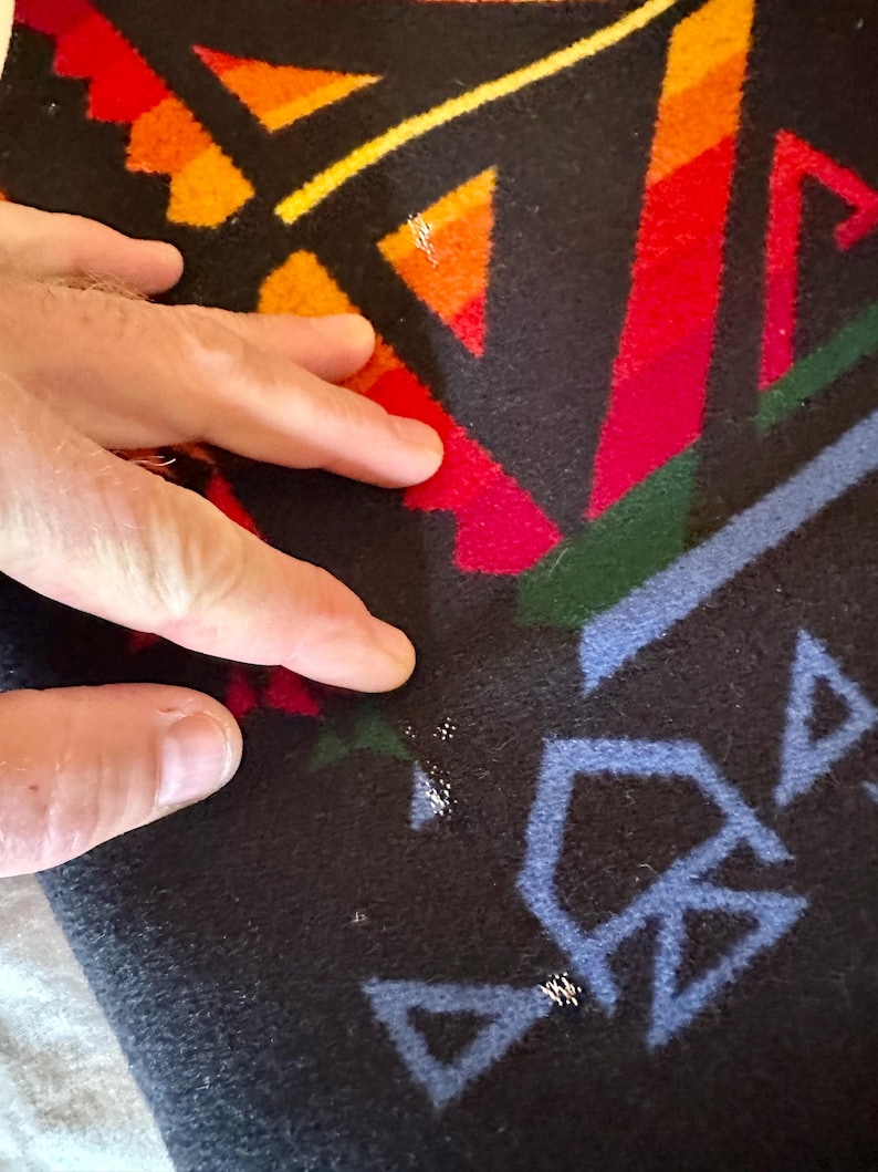 Puede incluir: Primer plano de un textil negro con un patr&oacute;n geom&eacute;trico en rojo, naranja, amarillo, verde y azul. El dise&ntilde;o presenta tri&aacute;ngulos y formas angulares. El textil tiene un aspecto suave y texturizado.
