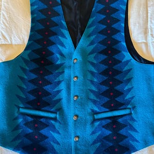 May include: A turquoise and navy blue patterned vest with a V-neck and four buttons. The vest features a geometric design with red accents and two front pockets. The interior lining is black.