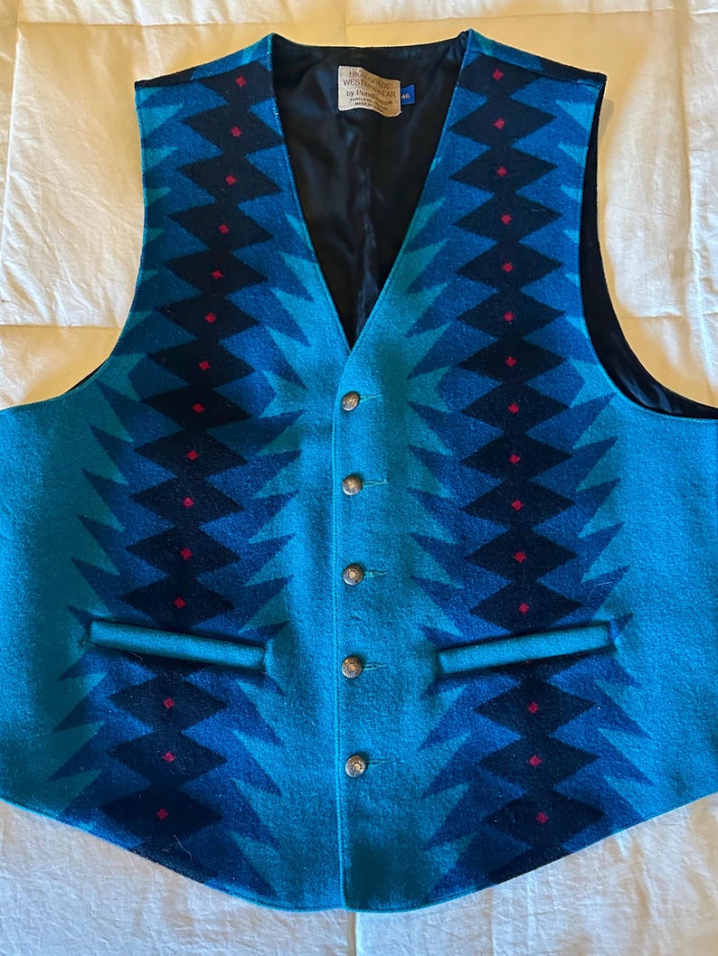 May include: A turquoise and navy blue patterned vest with a V-neck and four buttons. The vest features a geometric design with red accents and two front pockets. The interior lining is black.