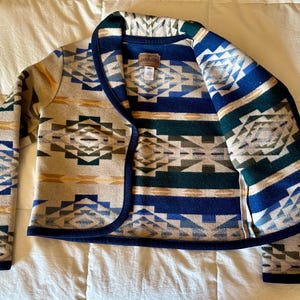 Może przedstawiać: Kurtka we wzory w odcieniach beżu, niebieskiego, zielonego i ż&oacute;łtego. Kurtka ma geometryczny wz&oacute;r i ciemnoniebieskie wykończenie. Kurtka z otwartym przodem wykonana jest z grubego, tkanego materiału.