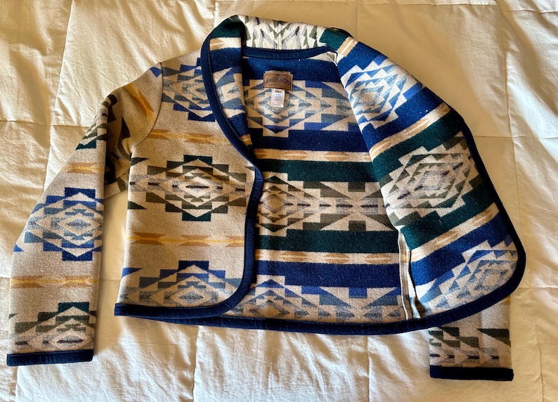 Może przedstawiać: Kurtka we wzory w odcieniach beżu, niebieskiego, zielonego i ż&oacute;łtego. Kurtka ma geometryczny wz&oacute;r i ciemnoniebieskie wykończenie. Kurtka z otwartym przodem wykonana jest z grubego, tkanego materiału.